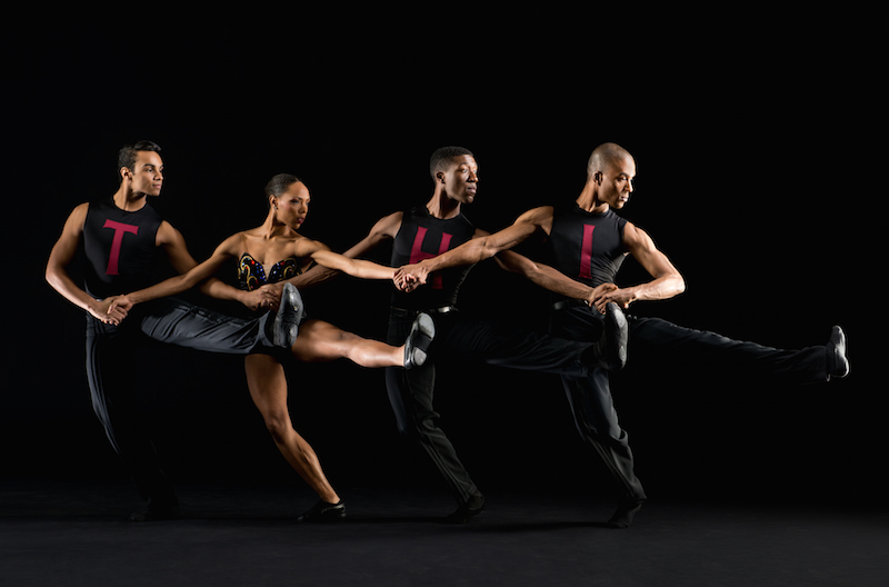 A quartet link arms and jut their left legs out as if they were doing a quirky kick line